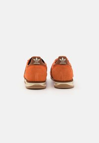 Oranžové semišové tenisky s hnědými akcenty, s texturovaným svrškem, polstrovaným límcem a gumovou podešví. Logo Adidas na patě.