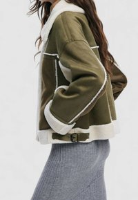 Olivgrön jacka med vita fårskinnsdetaljer, utformad med krage, framfickor och midjebälte. Kort, boxig passform med strukturerat tyg.