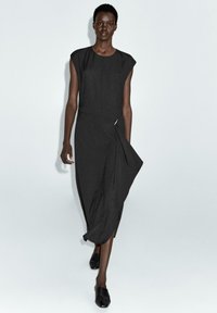 Schwarzes, ärmelloses Kleid mit strukturierter Silhouette, drapiertem Vorderteil und seitlichem Schlitz, aus einem glatten, leichten Stoff gefertigt.