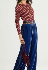 Burgunderfarbener Langarmpullover mit kreisförmigen Mustern, kombiniert mit weiten, königsblauen Samthosen. Akzente setzen ein Perlengürtel und eine kastanienbraune Beuteltasche mit Kordelzug.