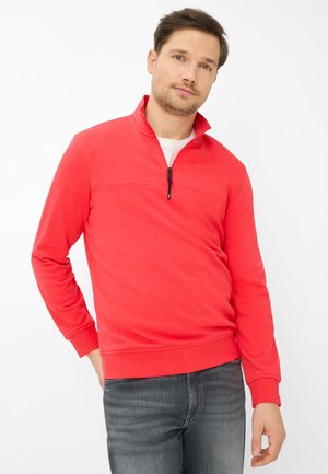 Uomo che indossa un maglione rosso con cerniera a mezza altezza sopra una camicia bianca, in piedi con una mano in tasca su uno sfondo bianco uniforme.