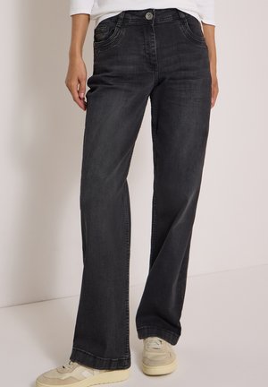 Person iført sorte jeans med lige ben, hvide sneakers og en hvid bluse med lange ærmer, stående mod en ensfarvet baggrund.
