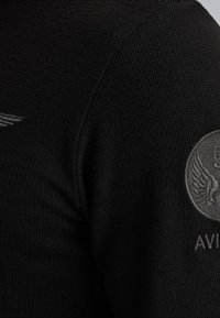 Schwarzer Strickpullover mit strukturierter Musterung, ausgestattet mit einem grauen gestickten Flügel-Emblem auf dem Ärmel und dem Text "AVIAN" darunter.