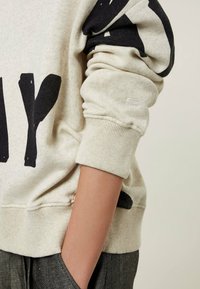 Světle šedá mikina s černým abstraktním potiskem. Hladké manžety a lem. Měkká, strukturovaná látka. Ruka modelu v kapse, oblékající šedé kalhoty s potiskem.
