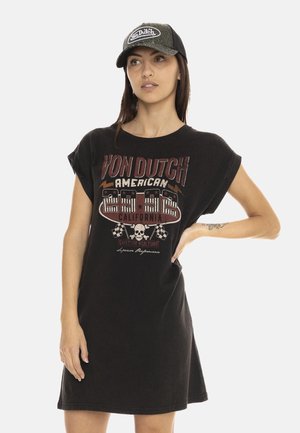 Femme portant une casquette noire Von Dutch et une robe noire oversize avec un texte graphique style vintage et un motif de crâne, posant avec une main sur la hanche.