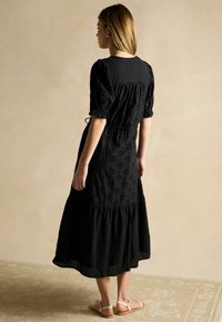 Schwarzes Kleid mit Puffärmel, strukturiertem Stoff, gestuftem Saum und floraler Stickerei. Kombiniert mit hellen Sandalen.