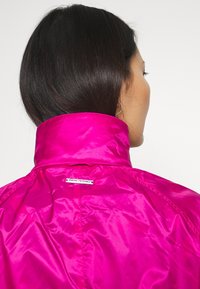 Žena s tmavými vlasy, oblečená v lesklé jasně růžové bundě s vysokým límcem, otočená zády, na kterých je vidět malý kovový štítek.