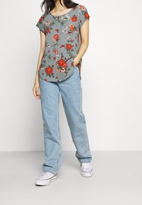 Top corto de manga corta en gris con patrones rojos y verdes; combinado con jeans de pierna recta azul claros y zapatillas blancas. Presenta bolsillos laterales.