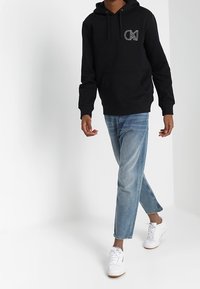 Felpa nera con cappuccio e tasca frontale, logo bianco sul petto, abbinata a jeans azzurri chiari e sneakers bianche. Tessuto morbido e vestibilità casual.
