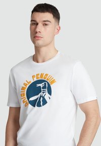 Weiße Baumwoll-T-Shirt mit einem runden Logo, einem blauen Kreis, einem Pinguin-Design und "Original Penguin" in orangefarbener Schrift. Kurze Ärmel.