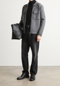 Šedá zipová bunda s dvěma předními kapsami, černý rolák, černé džíny a černé boty. Drží černou tašku. Minimalistický design.