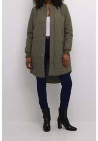 Oliwkowa pikowana długa kurtka z zatrzaskami i żeberkowymi mankietami, w połączeniu z ciemnoniebieskimi dżinsami i czarnymi botkami za kostkę.