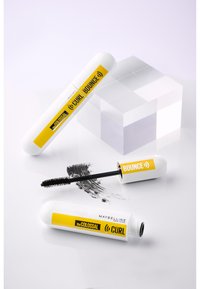 Mascara Maybelline avec un tube cylindrique blanc et des accents jaunes, présentant une texture en relief, un applicateur en brosse et une empreinte de mascara noir.