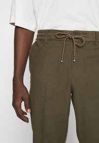 Personne à la peau foncée portant un pantalon vert olive avec une taille à cordon noué et une chemise blanche à manches courtes, main détendue sur le côté.