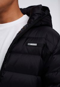 Zwarte donsjas met capuchon, voorzien van een zichtbaar logolabel en een getextureerd doorgestikt patroon. De binnenkant heeft een contrasterend ontwerp.