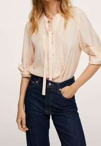 Blassrosa, gestreifte Bluse mit Knopfleiste, Halsbändern und hochgekrempelten Ärmeln; kombiniert mit dunkelblauen Jeans.