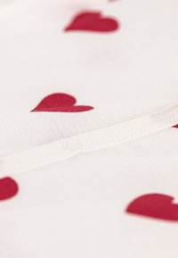 Λευκό ύφασμα με διάσπαρτα κόκκινα σχέδια καρδιάς και λεπτομέρεια κουμπιού στο κάτω μέρος. Απαλή υφή με ελαφριά ελαστικότητα.