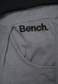 Szara tkanina o gładkiej fakturze z kieszenią. Na obszarze kieszeni wyszywane czarne logo "Bench." Prosty design.