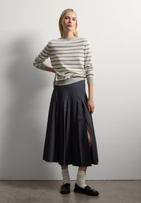 Jeune femme se tenant contre un mur, vêtue d'un pull rayé, d'une jupe midi plissée avec fente, de chaussettes blanches et de mocassins noirs.