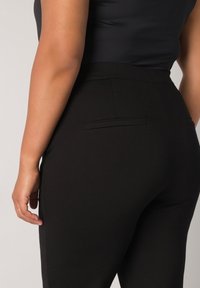 Zwarte op maat gemaakte broek met een hoge tailleband, voorzien van een gladde textuur en subtiele details op de achterzak. De aansluitende snit benadrukt de silhouet.