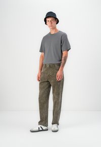 Gray short-sleeve t-shirt, olive green textured pants, black bucket hat, and white sneakers with black stripes. Turtle tattoo on arm.