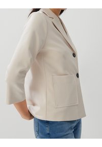 Světle béžový blazer s klopovým límcem, zapínání na jeden knoflík, tříčtvrteční rukávy a náprsní kapsou vpředu. Hladká textilie.