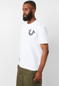 Camiseta de algodón blanca de manga corta, con un logo bordado en azul marino en el lado izquierdo del pecho, tejido suave y corte relajado.