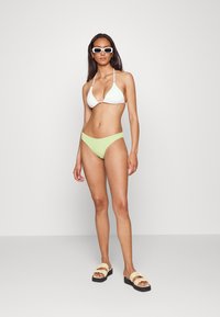 Top de bikini triângulo branco com textura canelada e alças ajustáveis, combinado com partes de baixo de bikini verde claro. A modelo usa óculos de sol brancos em forma de quadrado e sandálias confortáveis em tons de bege.