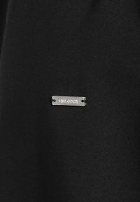Schwarzer Stoff mit glatter Textur, versehen mit einem silbernen Metallschild, auf dem "SMILODOX" steht, das seitlich angebracht ist und sich vom Hintergrund abhebt.