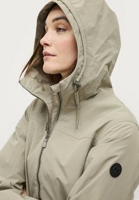 Lichtbeige waterdichte jas met een hoge kraag, verstelbare capuchon en ritssluiting. Bevat een rond zwart logo-opdruk op de mouw.