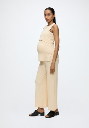 Femme enceinte debout de profil, portant un haut beige sans manches et un pantalon large, avec des sandales à talons noires sur un fond uni.