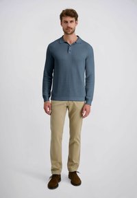 Mann in blauer gemusterter langärmliger Polohemd, beigen Hosen und braunen Schuhen steht vor einem schlichten hellen Hintergrund.