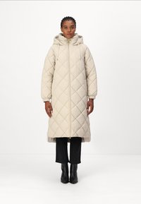 Beige gewatteerde jas met een capuchon, voorzien van een voorruit en elastische manchetten. Draag het over zwarte textuurbroek en enkellaarsjes.