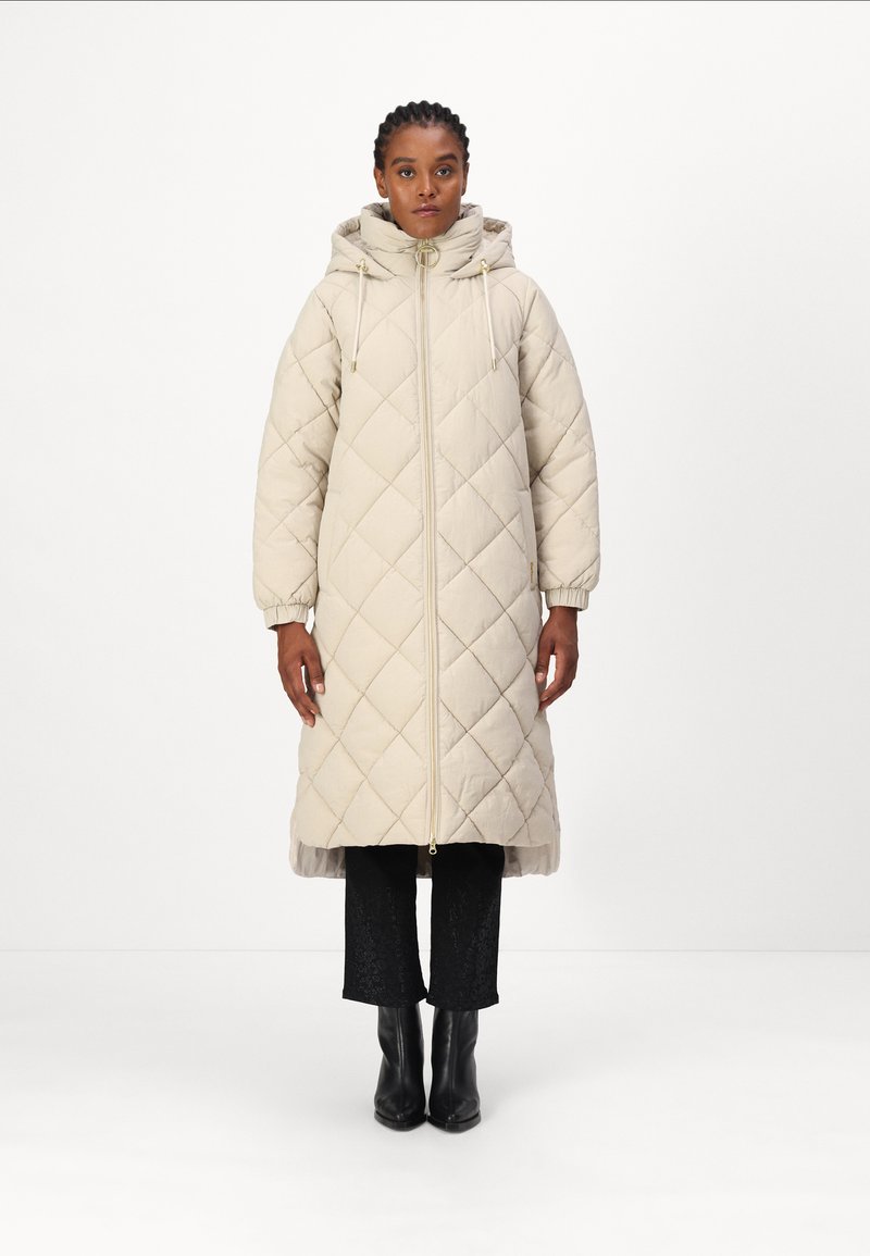Beige gewatteerde jas met een capuchon, voorzien van een voorruit en elastische manchetten. Draag het over zwarte textuurbroek en enkellaarsjes.