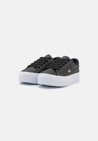 Zapatillas de cuero negro con una superficie texturizada, suela de goma blanca, cordones planos y detalle del logotipo en dorado en el costado y la lengüeta.