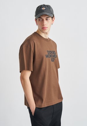 Mladý muž nosiaci hnedé tričko s krátkym rukávom s nápisom a tmavú čiapku, stojaci s rukami v vreckách, pozerajúci sa bokom na čistom pozadí.