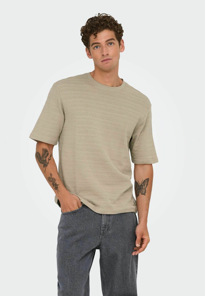 Lichtbeige katoenen t-shirt met korte mouwen en textuurpatroon, ronde halslijn, ontspannen pasvorm; gecombineerd met donkergrijze spijkerbroek en zichtbare armtatoeages.