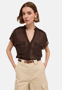 Transparente braune Kurzarmbluse mit zwei vorderen Taschen, Knopfverschluss und Schulterklappen, kombiniert mit beigen, hoch taillierten Hosen und einem dekorativen Gürtel.