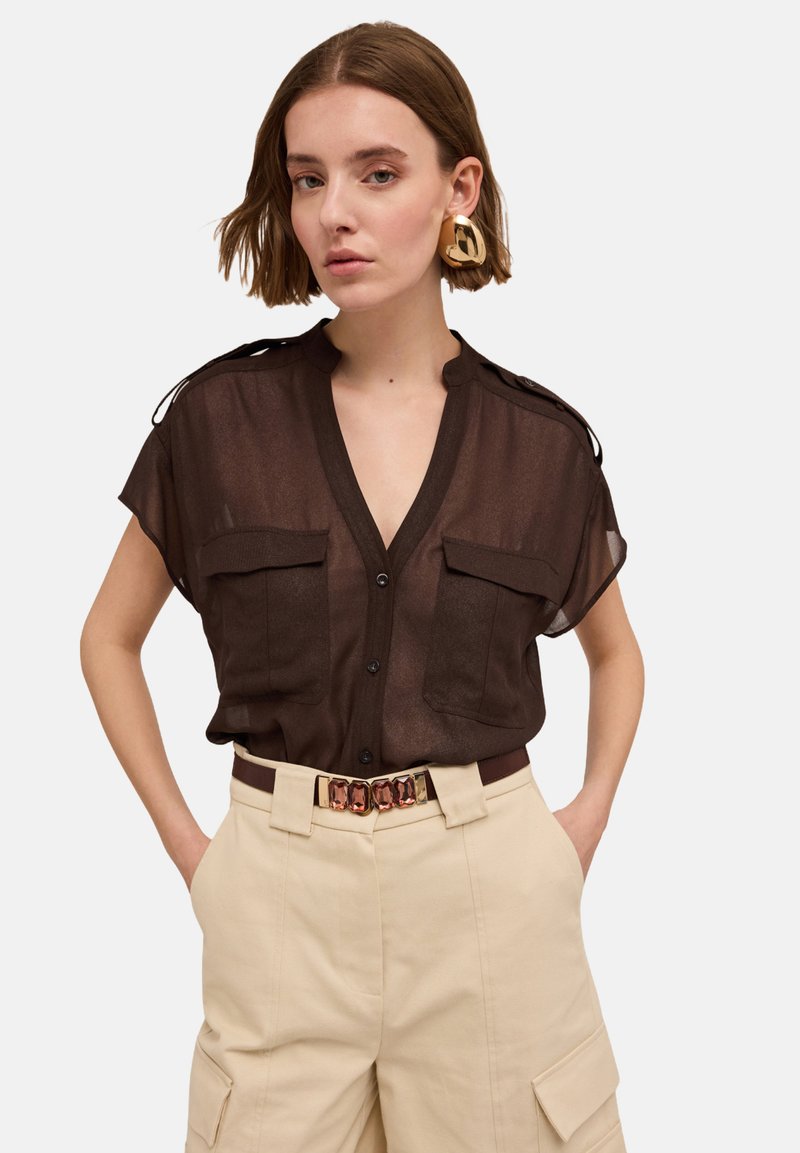 Transparente braune Kurzarmbluse mit zwei vorderen Taschen, Knopfverschluss und Schulterklappen, kombiniert mit beigen, hoch taillierten Hosen und einem dekorativen Gürtel.
