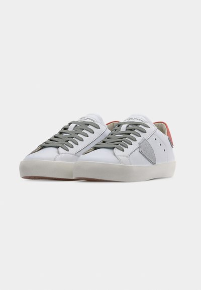 Baskets en cuir blanc avec lacets gris, un patch latéral gris et un détail orange au niveau du talon. Semelle en caoutchouc texturée. Design minimaliste.