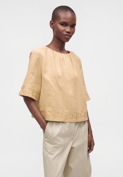 Femme aux cheveux courts portant un haut beige ample avec un ourlet et des manches brodés, associé à un pantalon beige clair, debout les mains dans les poches.
