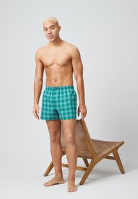 Groene geruite boxershorts met een elastische tailleband, met een mix van horizontale en verticale strepen. Model staat naast een houten stoel.