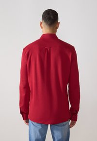 Camisa roja de manga larga con cuello clásico, costura en el yugo trasero y puños abotonados, combinada con jeans de color azul claro. Textura suave y ajuste entallado.
