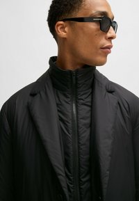 Giacca nera imbottita con colletto alto e chiusura con zip, caratterizzata da ampi revers e una texture liscia, abbinata a occhiali da sole neri eleganti.