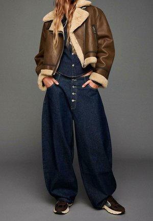 Femme portant une veste en peau de mouton marron, une chemise en jean boutonnée, un jean large foncé et des baskets marron, les mains dans les poches.