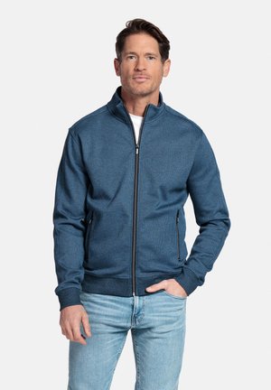 Homme portant une veste texturée bleue zippée et un jean bleu clair, debout avec une main dans la poche devant un fond uni.