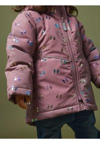 Barn iført en lyserød jakke med glitrende sommerfuglemønstre og denimbukser, stående foran en ensfarvet baggrund.