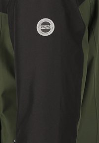 Svart och mörkgrön jacka gjord av slitstarkt material, med en rund silverlogotyp med texten "ZIGZAG" och rena linjer i designen.