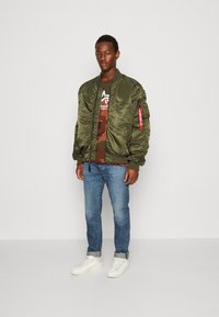 Olive green bomber jacket with ribbed cuffs, zip pockets, and red tag detail, paired with a camouflage t-shirt and blue jeans. White sneakers.