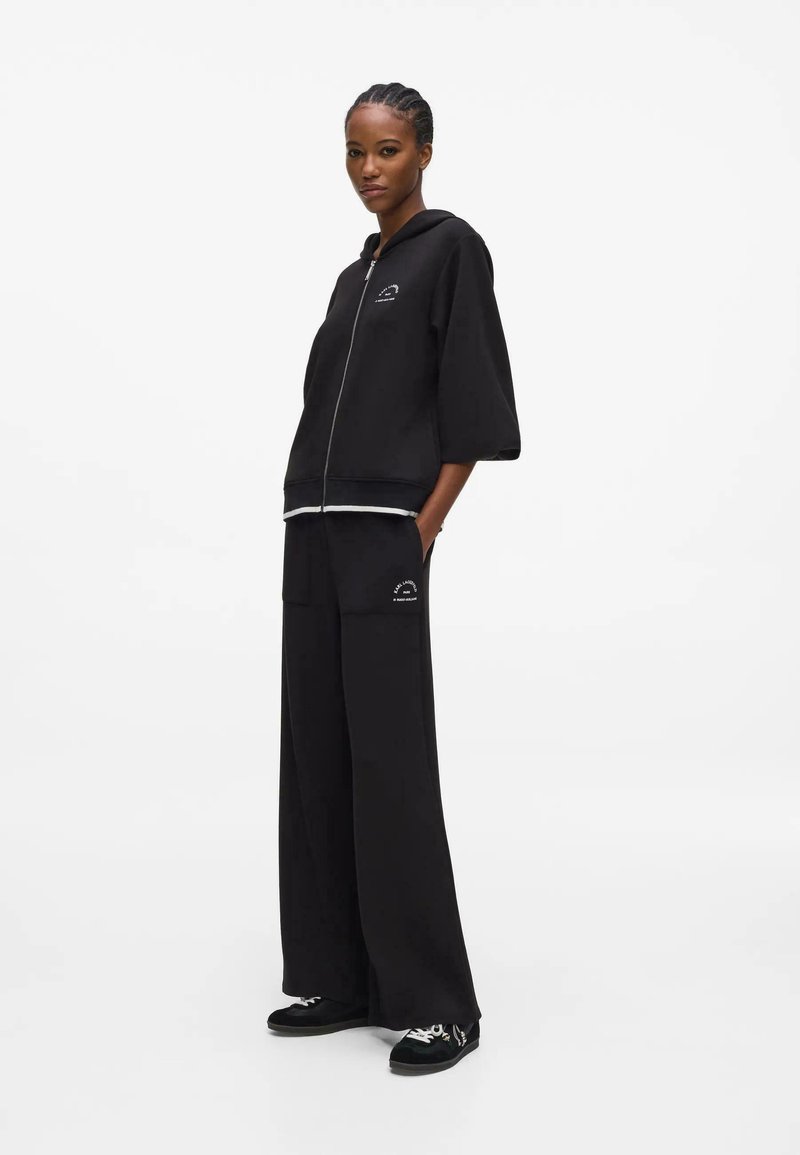 Femme debout portant un pantalon noir à jambes larges et un sweat à capuche noir zippé avec des baskets blanches sur fond blanc.